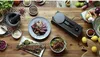 A top-down view of a wooden table with various food items and kitchen tools arranged, including a whole cooked fish, a plate of sliced meat, a bowl of noodles, and a mortar and pestle. Náhľad obrázku produktu