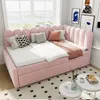 A pink upholstered daybed with multiple pillows and a throw blanket is set up in a bright room with a large window overlooking a body of water. Two small white tables with decorative items and books are placed in front of the daybed. Náhľad obrázku produktu