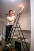 A woman is changing a lightbulb in a kitchen using a stepladder. Anteprima dell’immagine prodotto