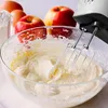 A hand mixer is used to mix ingredients in a glass bowl, with apples, eggs, and flour in the background. Produktbild-Vorschau