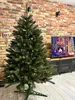A tall, artificial Christmas tree stands in a room with a brick wall and a TV in the background. Podgląd zdjęcia produktu
