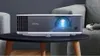 A BenQ projector is placed on a coffee table in a living room setting, with a couch and a game controller visible. Produktbild-Vorschau