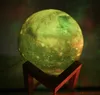 A moon-shaped lamp with a wooden stand, illuminated with green and yellow light, with some red and green light effects in the background. Náhľad obrázku produktu