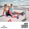 A couple sitting on a yoga mat on a rooftop, with weights and a yoga mat roller nearby. Produktbild-Vorschau