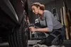 A woman is using a cordless impact wrench to tighten the lug nuts on a car wheel in a garage. Anteprima dell’immagine prodotto