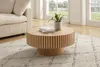 A round coffee table with a fluted design sits on a textured rug in a living room setting, next to a sofa. A small potted plant and books are on the table. Produktbild-Vorschau