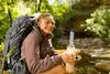 A woman with a large backpack is sitting outdoors, holding a water bottle with a filter. She is smiling and looking to the side. The background is a natural environment with trees and rocks. Podgląd zdjęcia produktu