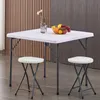 A white square table with two white stools in front of a wood-paneled wall. Produktbild-Vorschau