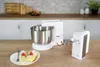 A white stand mixer with a silver bowl and a detached hand mixer attachment, next to a plate with two slices of cake topped with raspberries, on a wooden table. Anteprima dell’immagine prodotto
