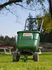 A green fertilizer spreader sits on a grassy lawn with trees and benches in the background. Anteprima dell’immagine prodotto