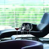 Four wine glasses filled with red and clear liquid are placed on a modern white table in front of a black lounge chair. The background features horizontal blinds with a blurred green outdoor scene visible through them. Produktbild-Vorschau