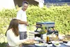 A couple is enjoying pizza outdoors with a pizza oven and a table full of food. Anteprima dell’immagine prodotto