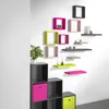 A collection of modular cube shelves in various colors mounted on a white wall, with a storage unit and a plant in the foreground. Produktbild-Vorschau