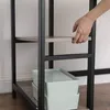 A person's hand is shown adjusting a shelf on a black metal framed shelving unit, with two light blue storage bins underneath. Náhled obrázku produktu