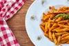 A plate of penne pasta with red sauce and a red chili pepper garnish, next to a red and white checkered napkin on a wooden table. Produktbild-Vorschau