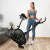 A woman in workout clothes stands next to a stationary bike in a home gym setting. Náhľad obrázku produktu