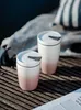 Two travel mugs with lids are placed on a wooden surface, with a blurred background of fabric and water. Voir les images du produit