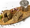 A round of pâté with a slice cut out, a slice of bread spread with pâté, garlic, peppercorns, and a stainless steel container on a wooden board. Podgląd zdjęcia produktu