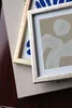 Two framed pictures are stacked on top of a book. The frames are a light wood color and a distressed white color. The pictures inside the frames are abstract art with blue shapes and line drawings. Produktbild-Vorschau
