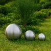 Three brushed stainless steel decorative spheres of varying sizes are placed on a grassy lawn in front of a green shrub. Podgląd zdjęcia produktu