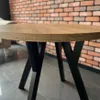 A close-up shot of a round wooden table with black criss-cross legs, set against a brick wall background. Voir les images du produit