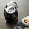 A black electric kettle with a silver handle is placed on a gray surface next to a muffin and some dried flowers. Produktbild-Vorschau