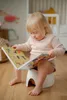 A young child with blonde hair and Down syndrome is sitting on a white potty and reading a book with colorful illustrations. Náhľad obrázku produktu