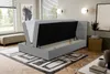 A bedroom scene with a grey boxspring bed with its storage compartment open, revealing the mattress and storage space. The room has a window, blinds, wall art, and a rug. Voir les images du produit