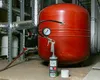 A red expansion vessel with a filling gun and pressure gauge attached, in a utility room with pipes and other equipment. Produktbild-Vorschau