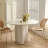 A round white dining table with a fluted base, set with two apples, a glass of water, and a vase of yellow flowers. Two chairs with wooden seats and metal legs are placed on either side of the table. A white folding screen is visible in the background. Produktbild-Vorschau