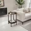 A round, two-tiered side table with a wood-grain top and black metal legs is placed next to a white sofa in a living room. Náhled obrázku produktu