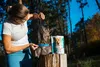 A woman is feeding a dog in a forest setting, with a bag of dog supplement next to them. Produktbild-Vorschau