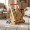 A wooden baby walker with various interactive toys is shown on a carpeted floor in a room with a sofa and shelves in the background. Produktbild-Vorschau
