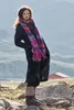 A woman wearing a dark coat and a colorful scarf stands in a rocky, outdoor landscape. Produktbild-Vorschau