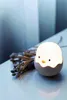 A small, egg-shaped light with a cute chick face, placed next to dried plant stems. Produktbild-Vorschau