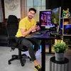 A man is sitting in a gaming chair at a desk with a computer and gaming accessories. Produktbild-Vorschau
