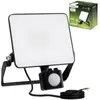 A black LED floodlight with a motion sensor is shown in front of a white background, with its packaging visible in the upper right corner. Náhled obrázku produktu
