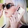 A woman wearing sunglasses holds a pink phone case with a clear back and a magnetic ring, while adjusting her sunglasses. Produktbild-Vorschau