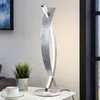 A modern silver LED table lamp is displayed on a white table next to a potted plant and a cup of coffee. Produktbild-Vorschau