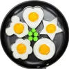 Five different shaped egg molds with fried eggs inside, placed in a black frying pan. There are also some green candies scattered around. Produktbild-Vorschau