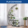A black metal plant stand with multiple shelves, holding various potted plants, placed indoors in front of sheer white curtains. Produktbild-Vorschau