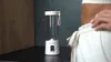 A portable blender sits on a kitchen counter next to a person's lower body. Podgląd zdjęcia produktu