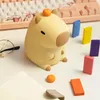 A cute capybara-shaped night light is placed on a desk next to a notebook, glasses, and colorful blocks, with a keyboard in the background. Podgląd zdjęcia produktu