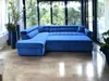 A large blue sectional sofa is shown in a sunlit room with large windows overlooking greenery. Náhled obrázku produktu