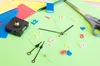 A collection of clock parts, including a clock movement, hands, and numbers, laid out on a green surface with some colored paper scraps. Anteprima dell’immagine prodotto