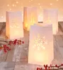 Four white paper lanterns with cut-out winter motifs are arranged on a wooden surface, with red berries in the foreground and blurred lights in the background. Produktbild-Vorschau