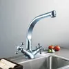 A chrome kitchen faucet with two handles is shown in a sink, with tomatoes and limes on the counter. Náhled obrázku produktu