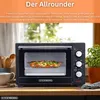 A black mini oven with food inside, placed on a wooden countertop with a window and plants in the background. Podgląd zdjęcia produktu