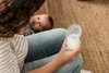 A person is holding a breast pump with milk in it, with a baby lying on their lap. Náhled obrázku produktu