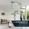 A woman is hanging laundry on a ceiling-mounted electric clothes dryer in a modern, well-lit room with large windows. Produktbild-Vorschau